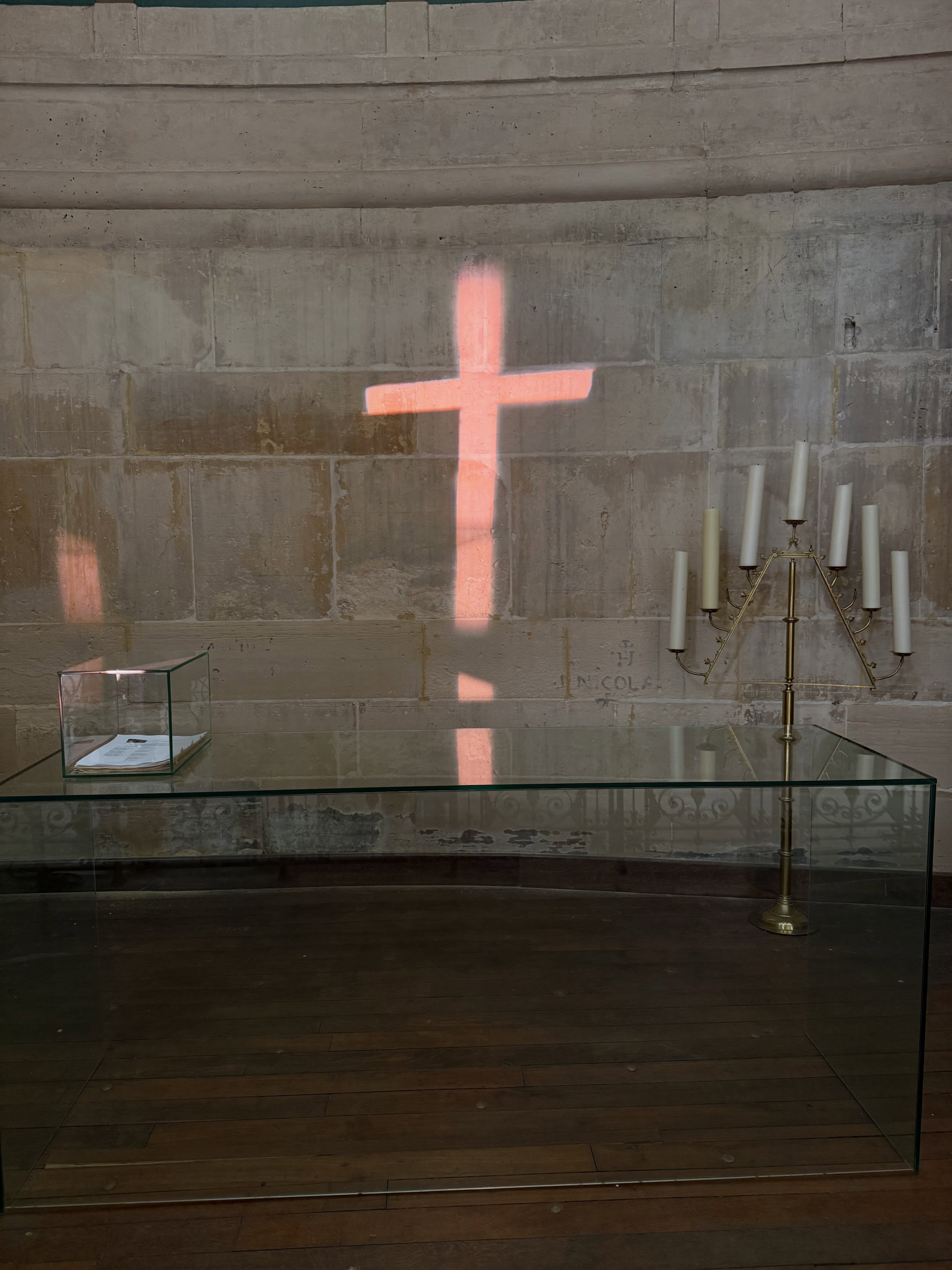 A pink cross-shaped reflection falls across a concrete church wall behind a dark altar.