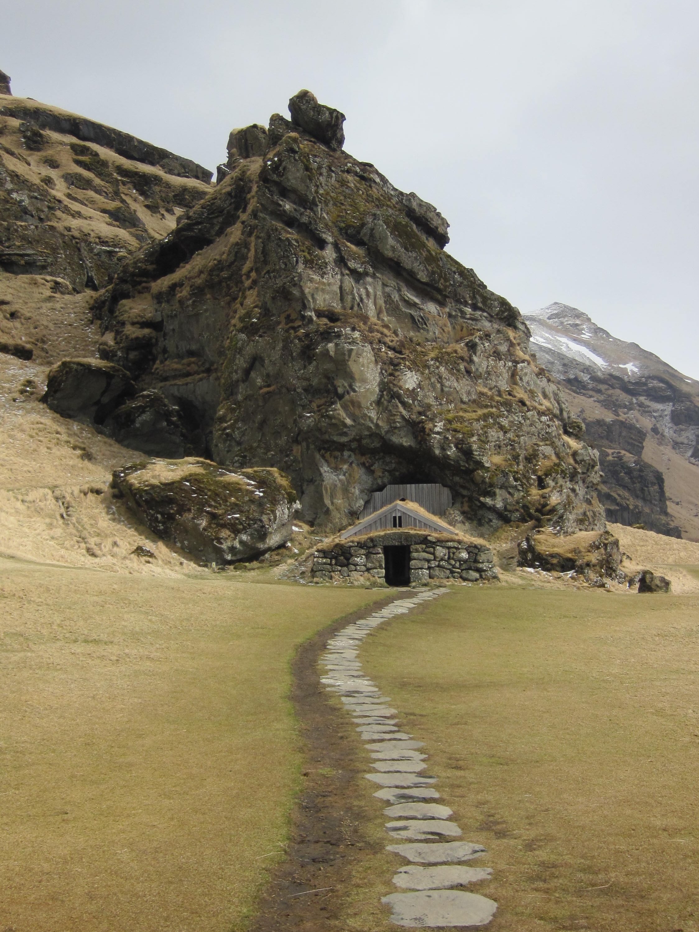 A stone path leads toward a small turf-roof building beneath a rocky Icelandic hillside.